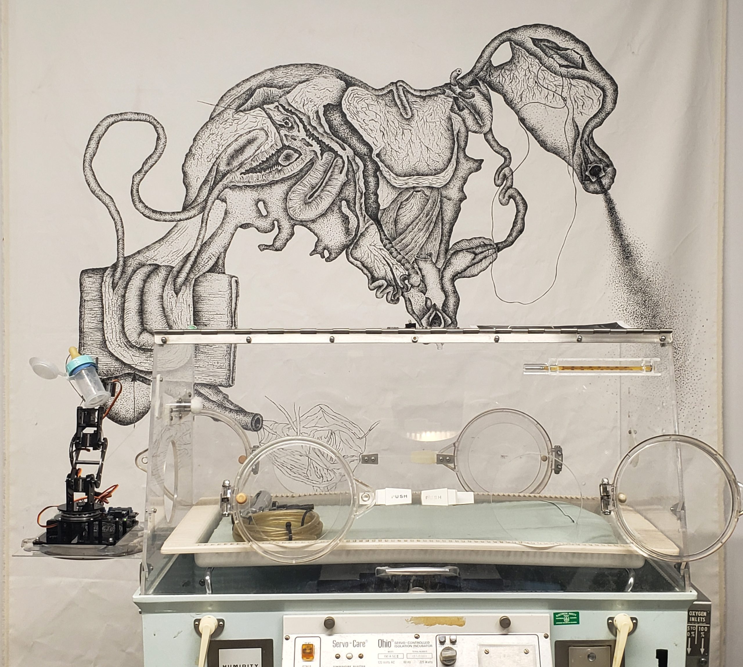 "Bio Box" installation image, a photo of an incubator with a robotic arm and background drawing of chalkboard paint on canvas.